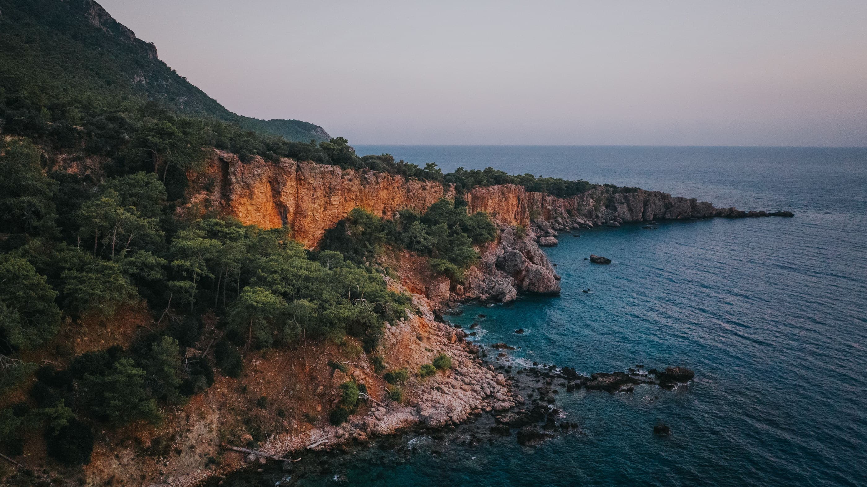 A cliff headland near Antalya at first light — pine and scrub running down warm limestone to deep blue Mediterranean water.