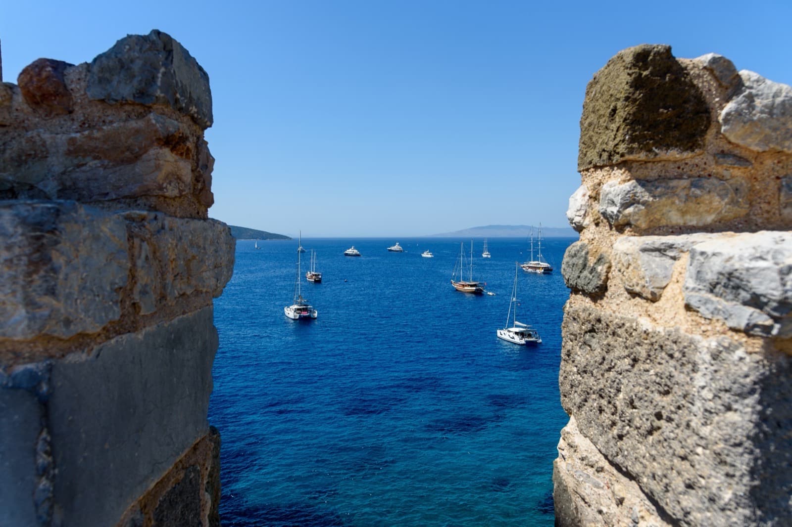 The Aegean seen between two weathered stone battlements of Bodrum castle, with sailing boats at anchor.