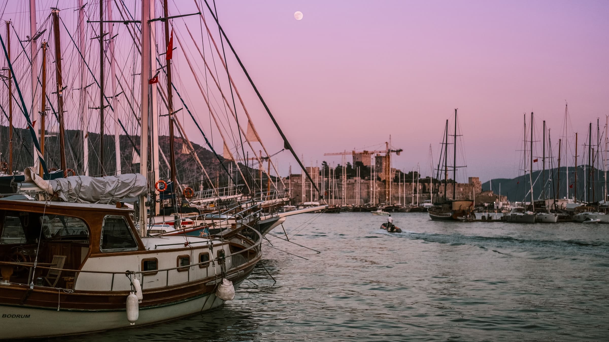 Wooden gulets moored along the Bodrum waterfront at dusk, masts against a pink sky with the castle and the moon behind.