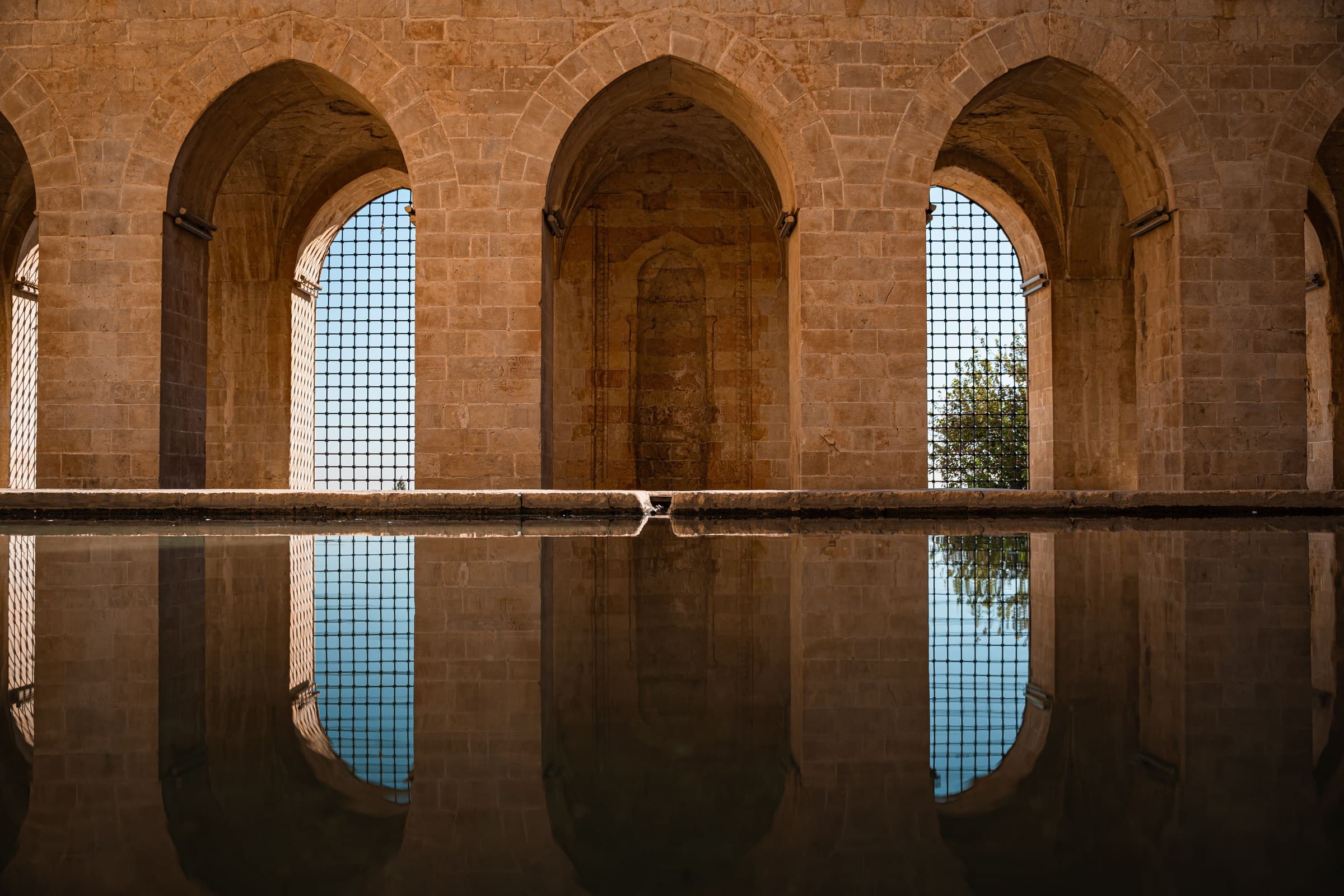 A limestone arcade in Mardin standing over a still pool, its pointed arches and the sky behind them reflected in the water.