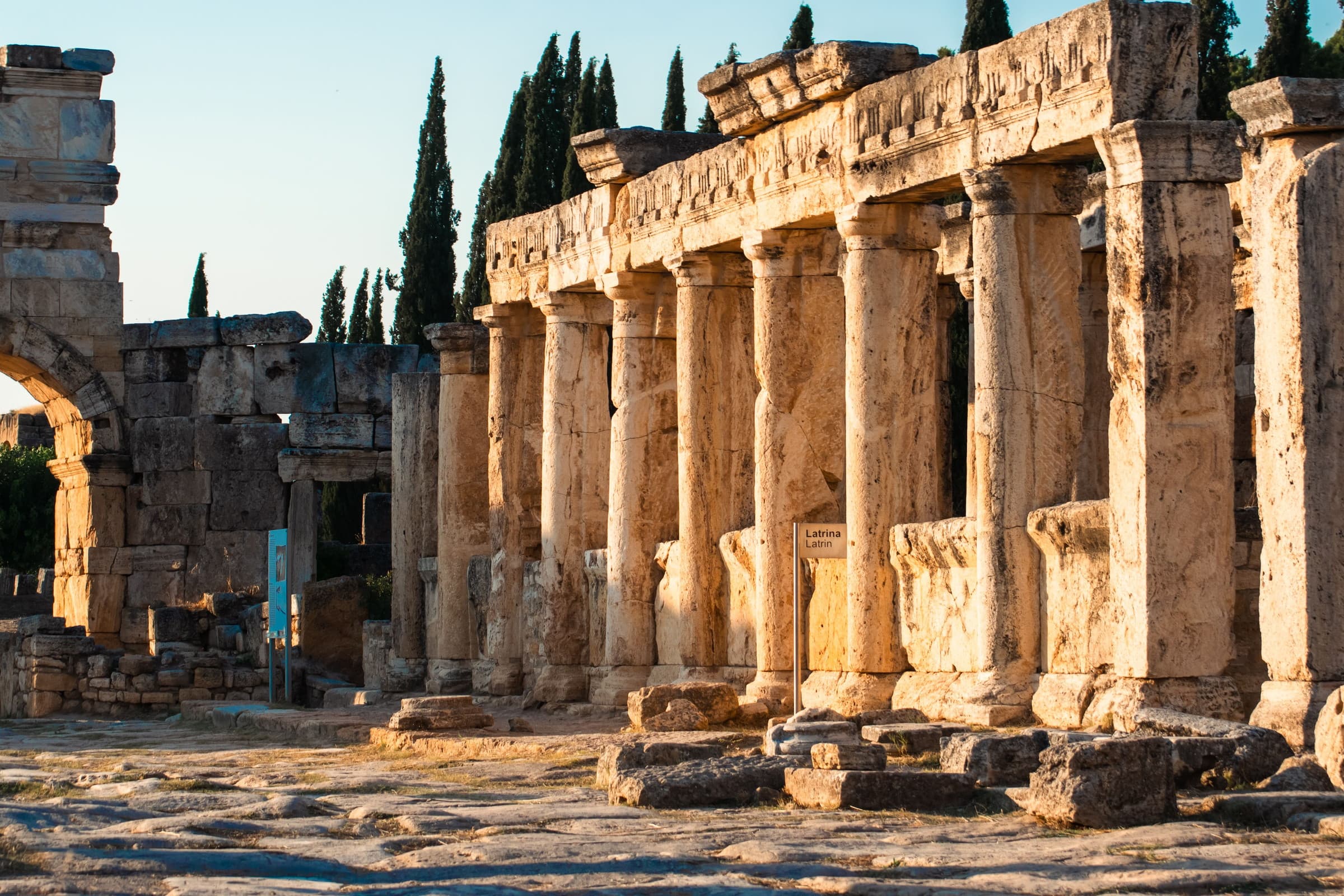 The colonnade at Hierapolis in low sun — limestone columns, a standing arch and cypresses along the ancient street.
