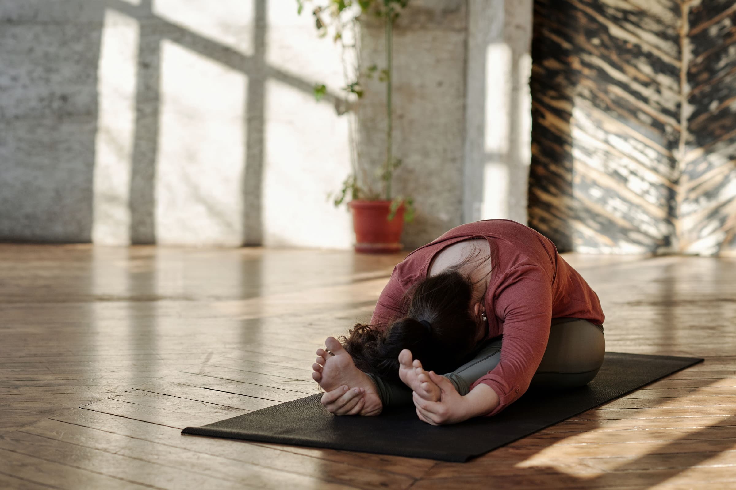 A person folded forward over crossed legs on a mat, in a warm room of textured plaster, timber and plants.